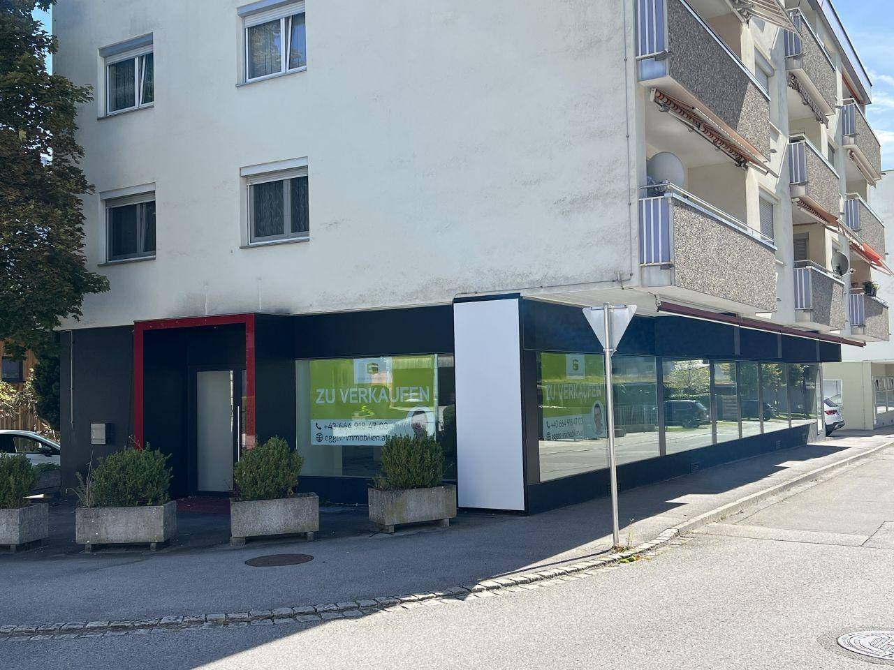 Eingangsbereich eines Geschäfts im Erdgeschoss eines Gebäudes mit großen Schaufenstern und Pflanzenkübeln.