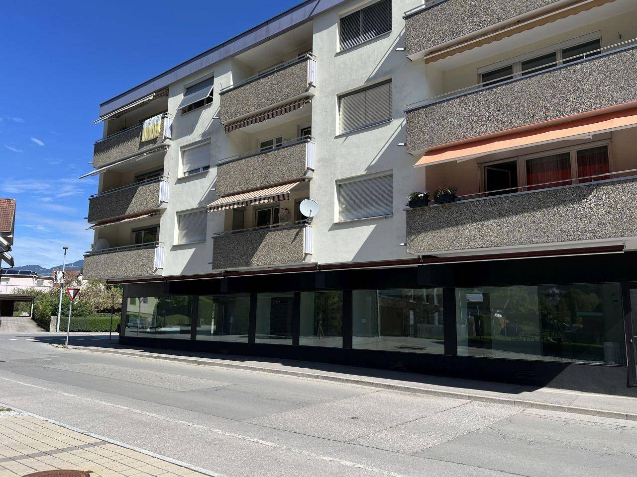 Ansicht eines Mehrfamilienhauses mit Balkonen und Geschäftsräumen im Erdgeschoss an einer sonnigen Straße.