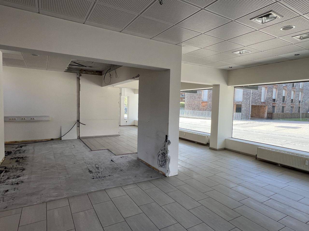 Geräumiger Ladenbereich im Rohbauzustand mit Fliesenboden, abgehängter Decke und großen Schaufenstern.