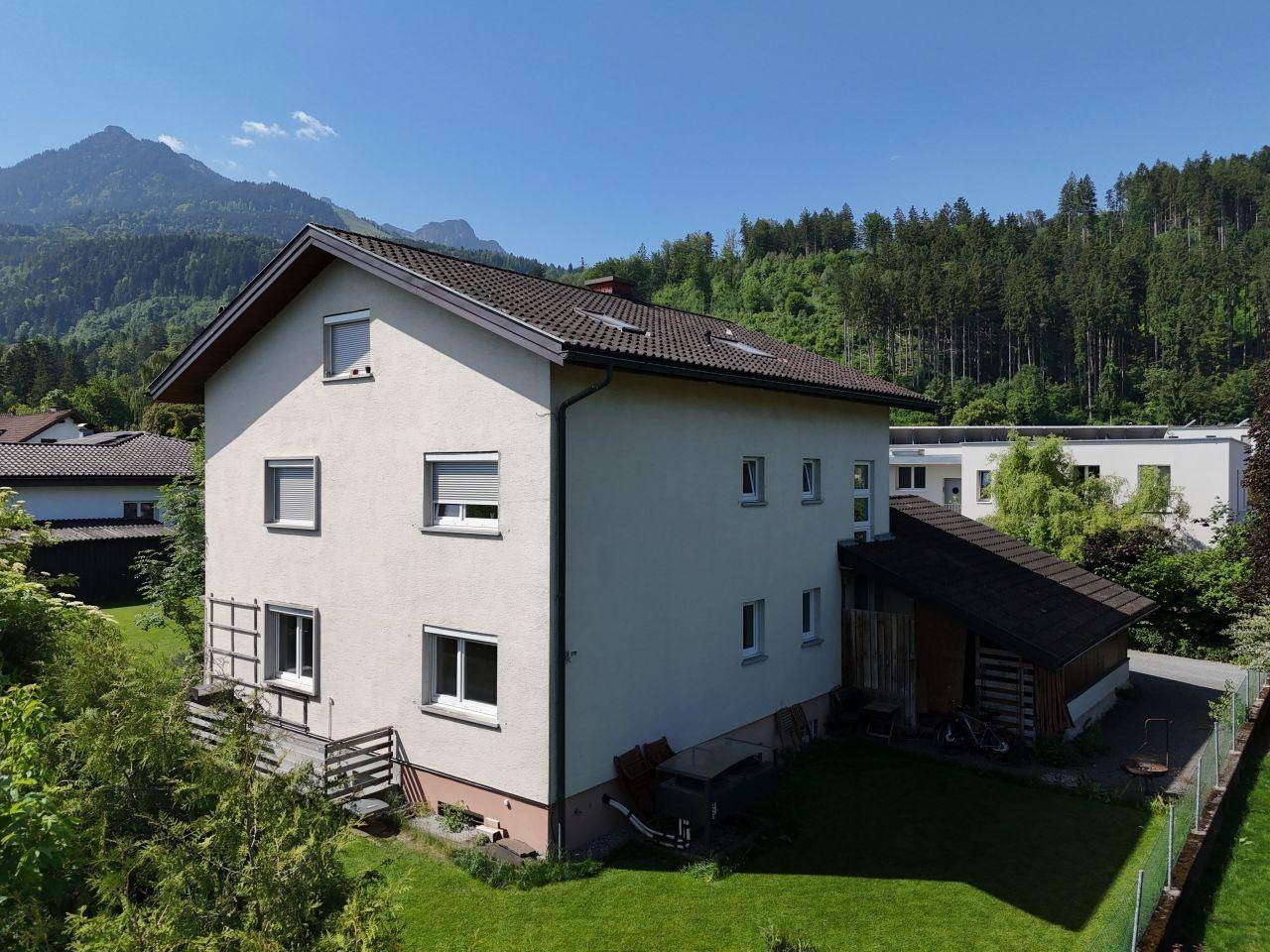 Ansicht des Hauses von der Seite mit gepflegtem Garten und bewaldeten Bergen im Hintergrund.