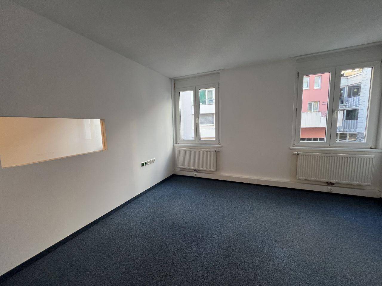 Geräumiger Büroraum mit Teppichboden und zwei Fenstern für ausreichend Tageslicht.