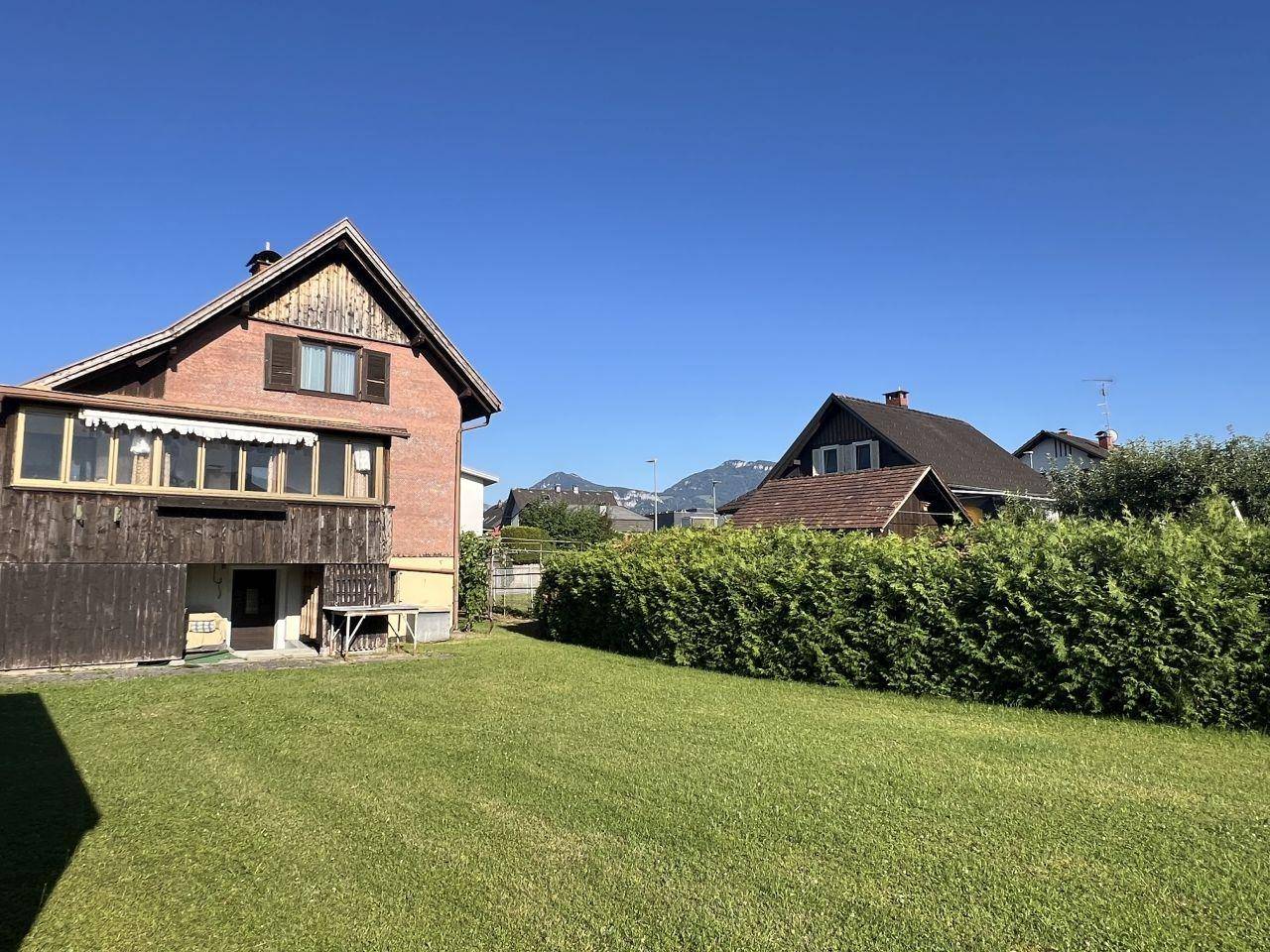 Hinteransicht eines Hauses mit Holzanbau und großem Garten, umgeben von Hecken und mit Bergblick.