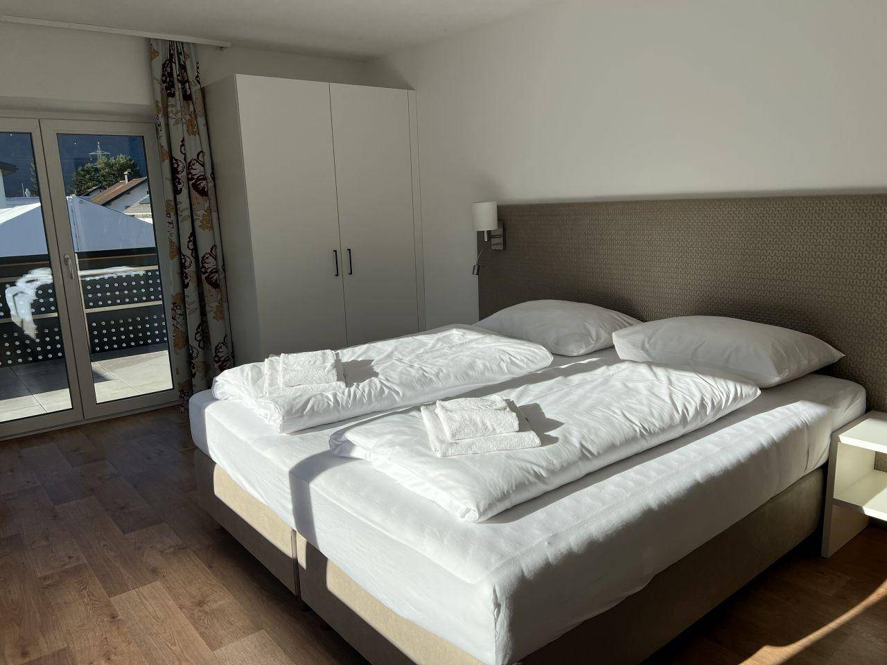 Sonnenverwöhntes Schlafzimmer mit Doppelbett, weißem Kleiderschrank und direktem Balkonzugang.