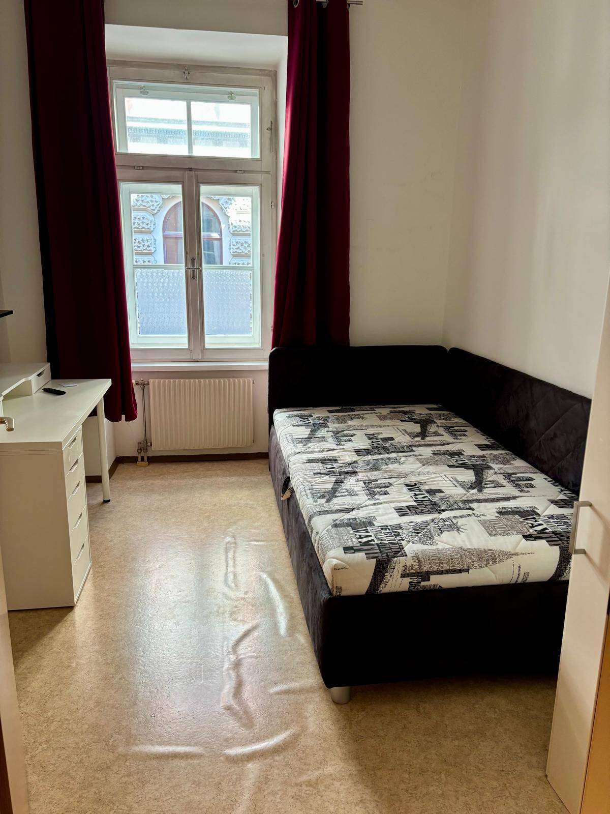 Helles Schlafzimmer mit einem Einzelbett, weißem Schreibtisch und Fenster mit roten Vorhängen.