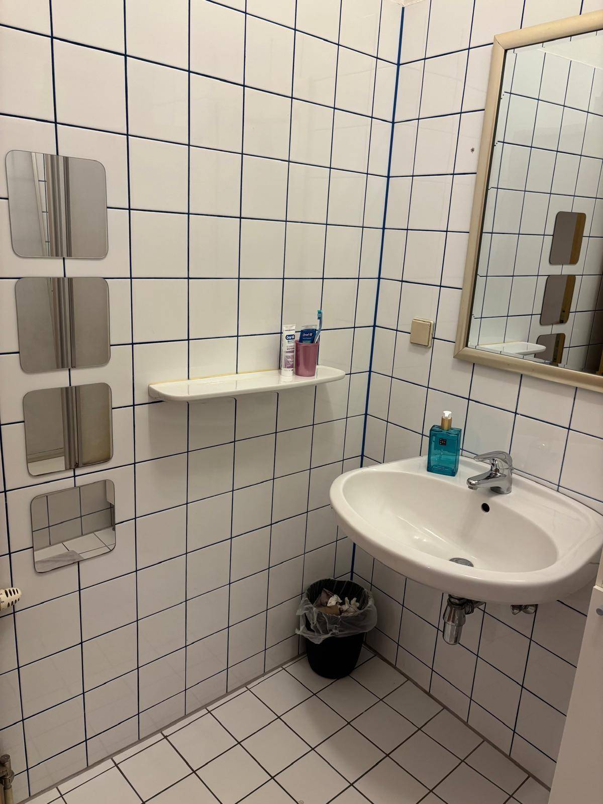 Badezimmer mit weißen Fliesen, einem Waschbecken und einem Spiegel, typisch für die 80er Jahre.