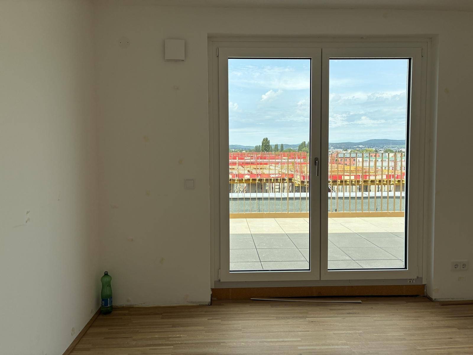 Geräumiges Zimmer mit Holzboden und großer Fenstertür, die auf eine Baustelle blickt.