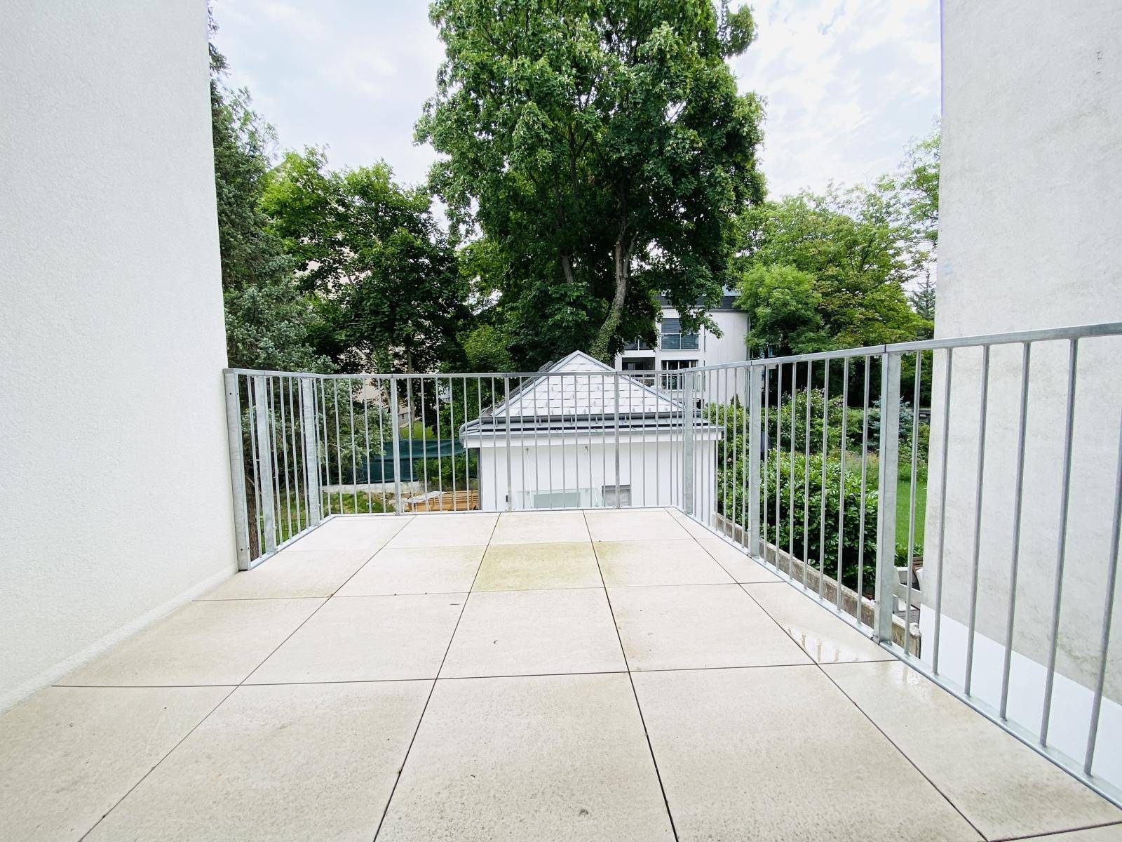 Geräumiger Balkon mit Fliesenboden und Metallgeländer, umgeben von Bäumen.