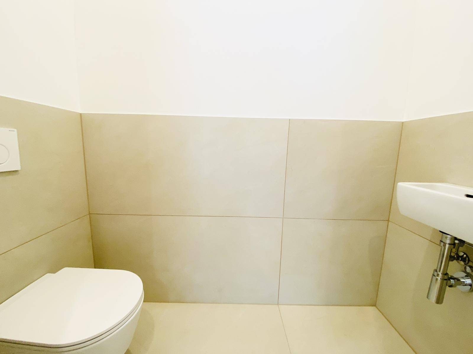 Kompaktes Gäste-WC mit beigefarbenen Fliesen, wandhängender Toilette und kleinem Waschbecken.