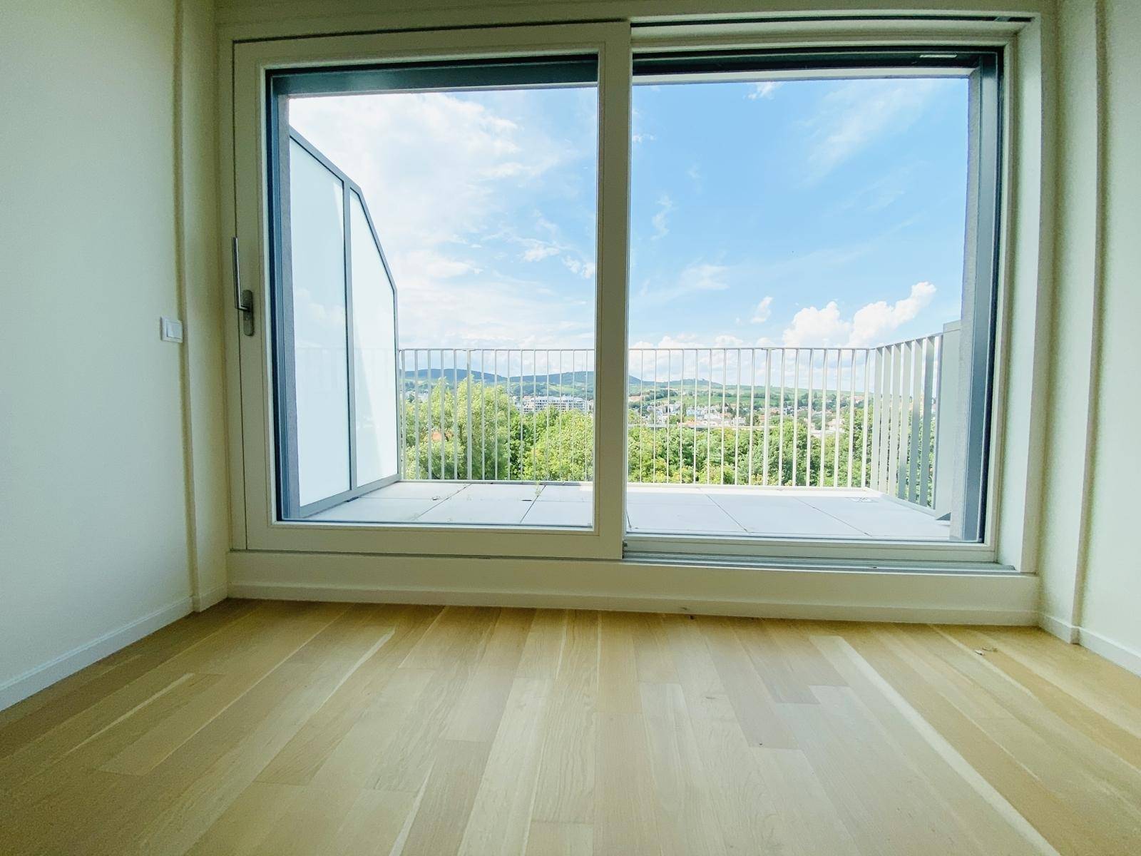 Heller Raum mit Holzboden und großer Schiebetür, die auf einen Balkon mit Panoramablick führt.
