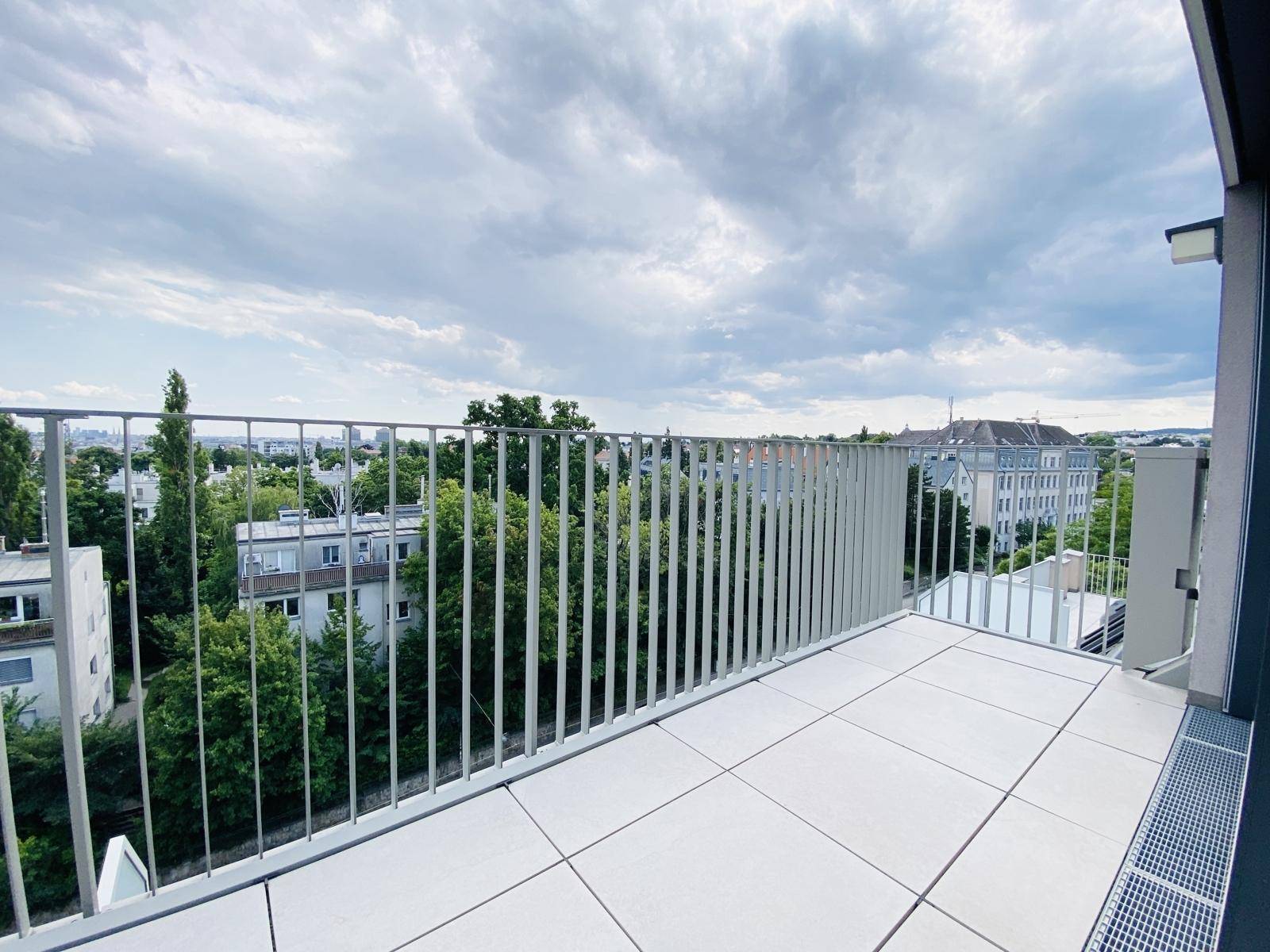 Balkon mit Fliesenboden und Metallgeländer, bietet einen Blick auf die Stadt und umliegende Bäume.
