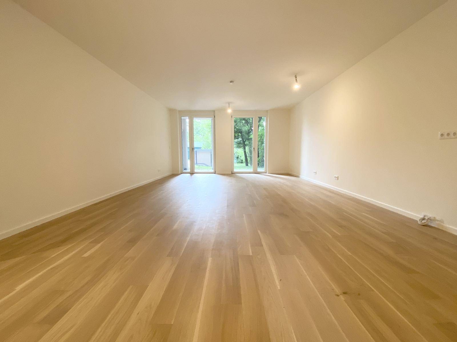 Weitläufiges Wohnzimmer mit hellem Parkett und viel Tageslicht durch die großen Fenster.