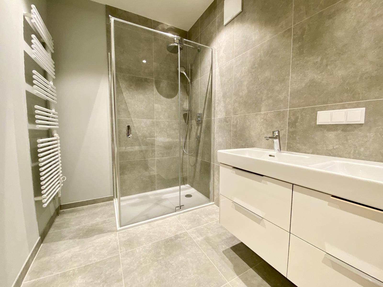 Modernes Badezimmer mit grauen Fliesen, Glasdusche, Doppelwaschbecken und Handtuchheizkörper.