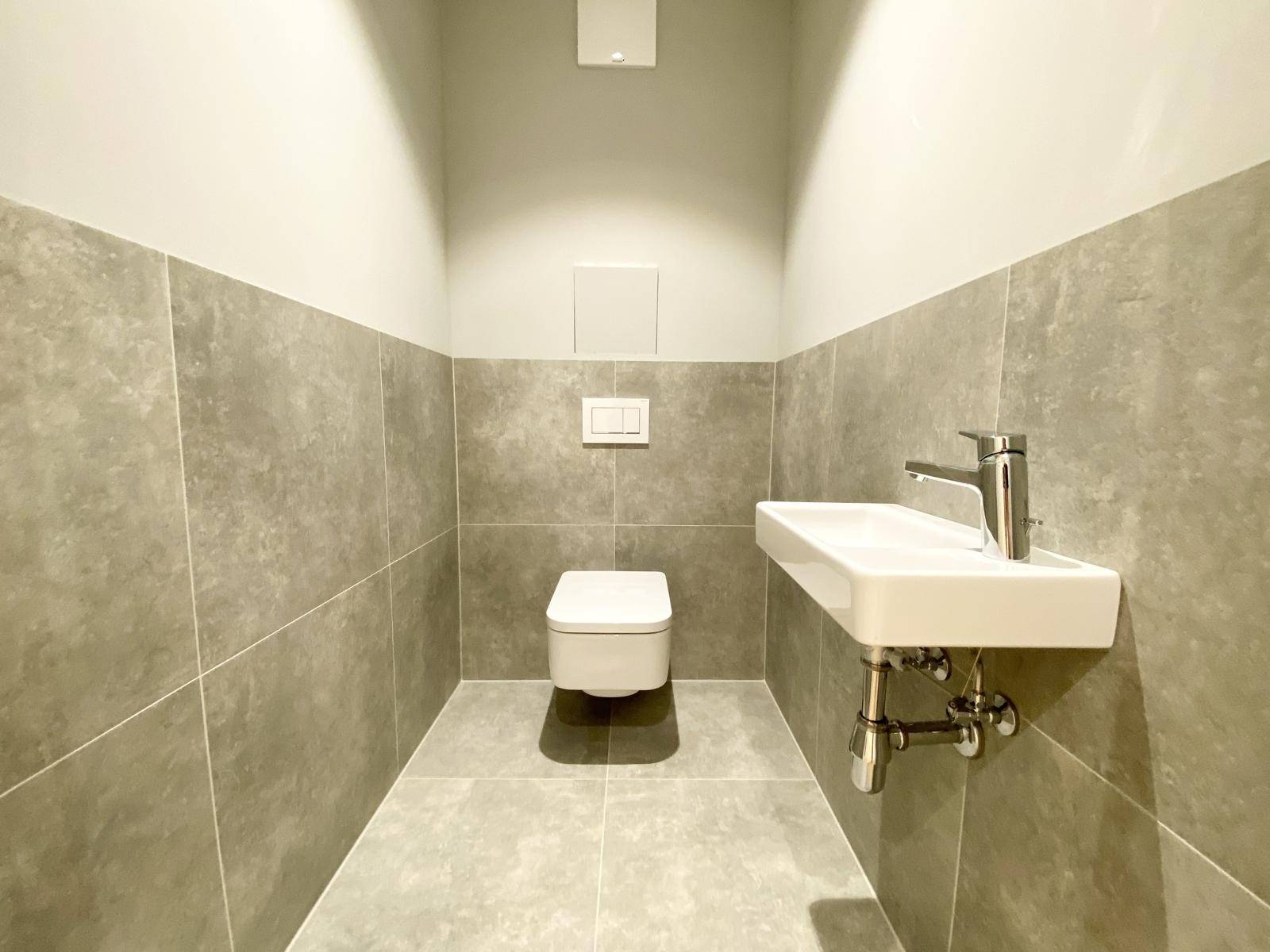Modernes Gäste-WC mit grauen Fliesen, wandhängendem WC und kleinem Handwaschbecken.