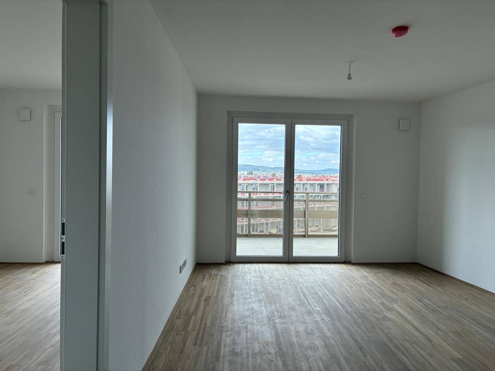 Heller Raum mit Holzboden und großem Fenster, das viel Tageslicht hereinlässt und Zugang zum Balkon bietet.