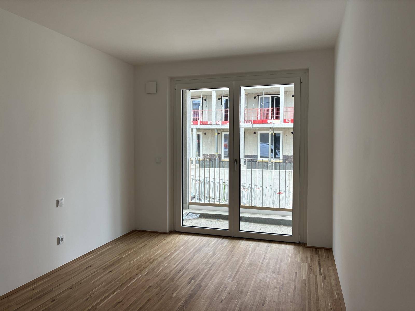 Helles Zimmer mit Holzboden und großem Fenster, das Zugang zu einem Außenbereich bietet.