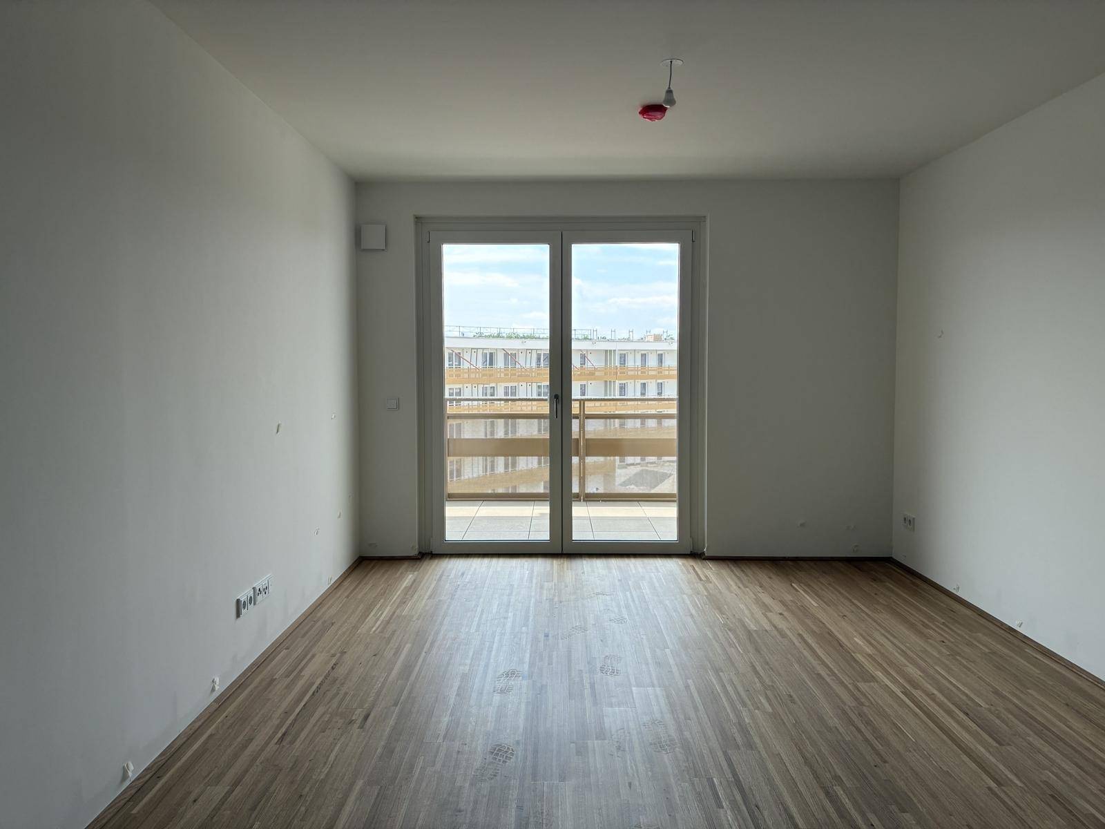 Helles, leeres Zimmer mit Holzboden und Zugang zu einem Balkon in einem Neubau.