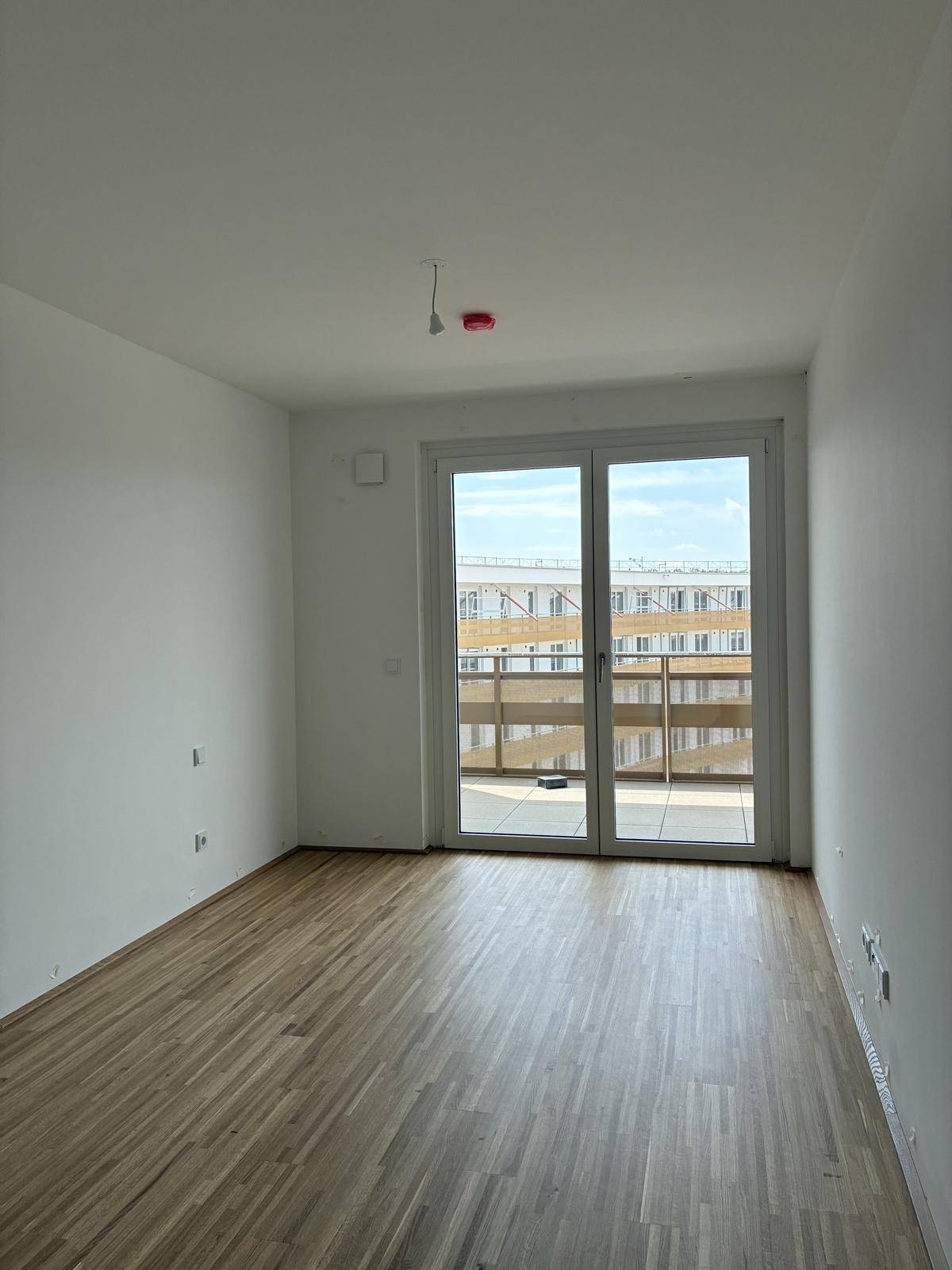 Geräumiges, helles Zimmer mit Holzboden und breitem Zugang zu einem Balkon.