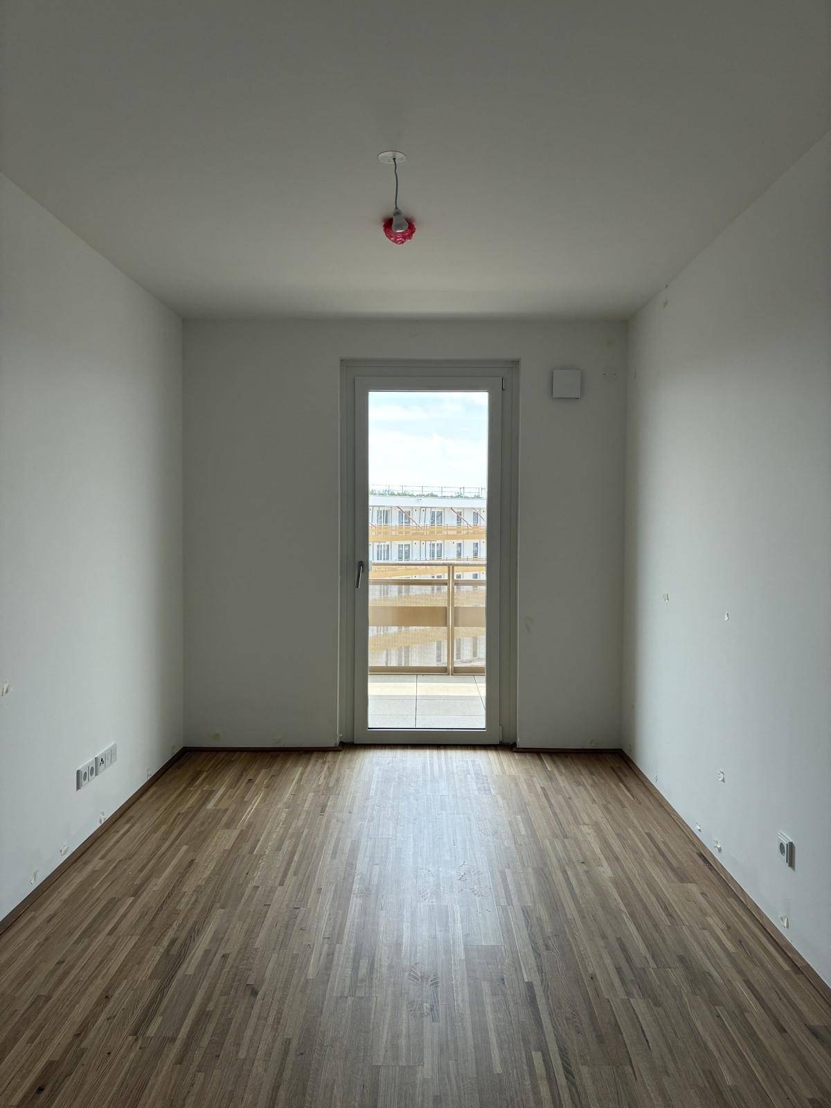 Leeres Zimmer mit Holzboden und Zugang zu einem kleinen Balkon, ideal als Schlafzimmer.