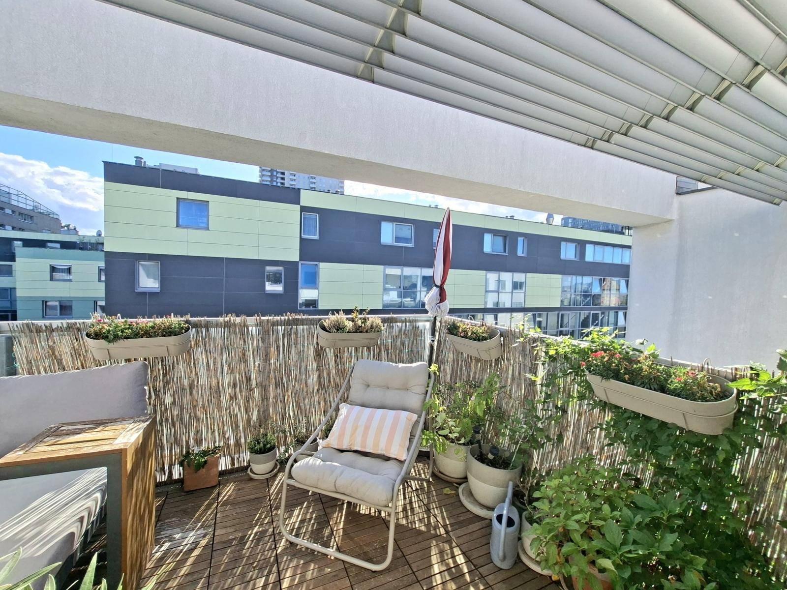 Gemütlicher Balkon mit Holzboden, Pflanzen und Sitzgelegenheiten, umgeben von Stadtgebäuden.