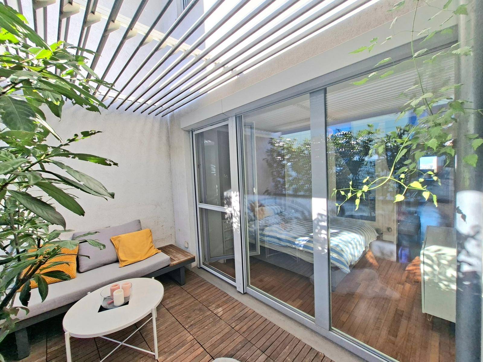 Gemütliche Terrasse mit Sitzgelegenheiten, Pflanzen und Blick in ein Schlafzimmer durch Glasschiebetüren.