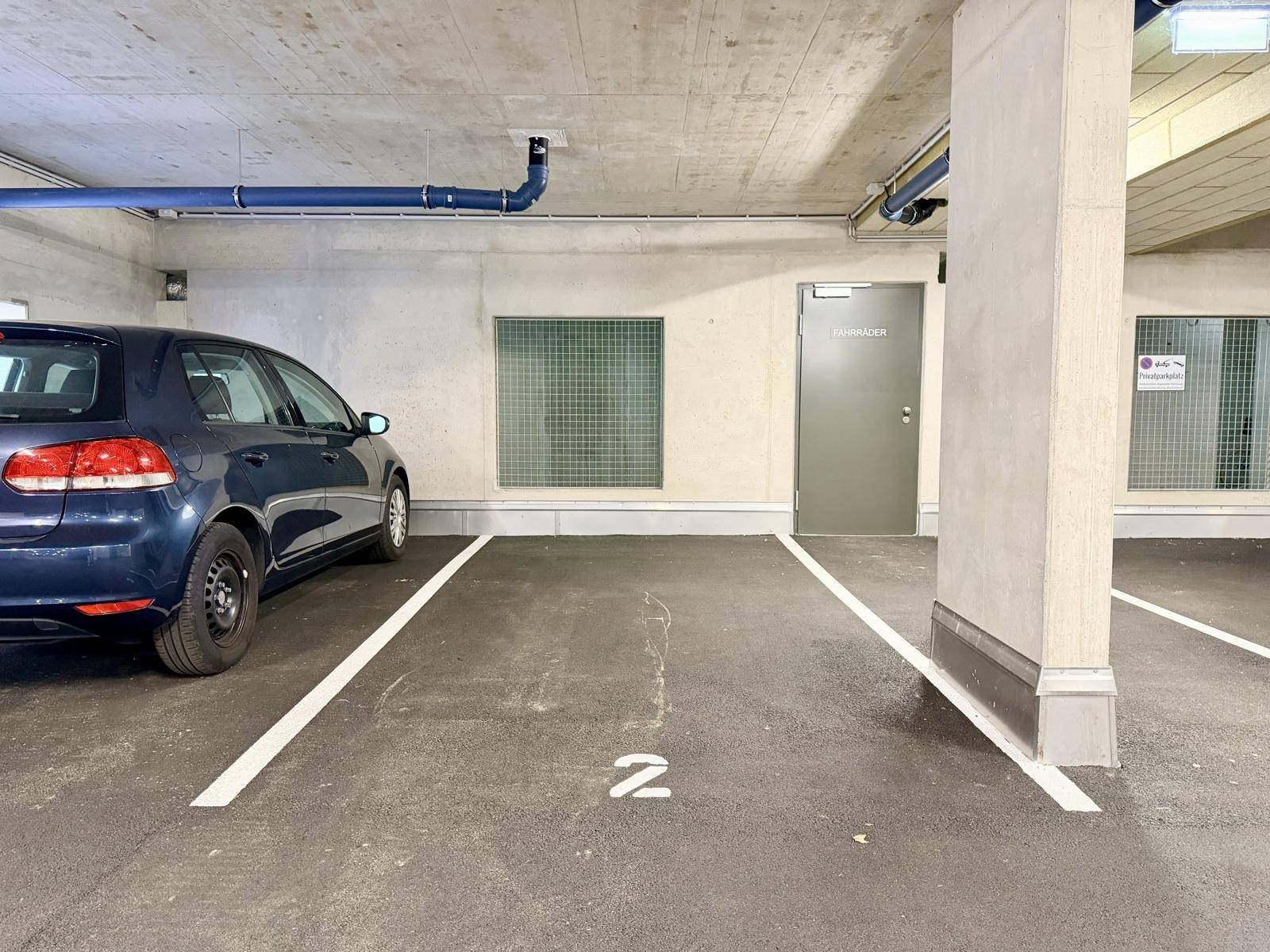 Einzelner Stellplatz in einer sauberen Tiefgarage mit Betonwänden, blauen Rohren und Beleuchtung.
