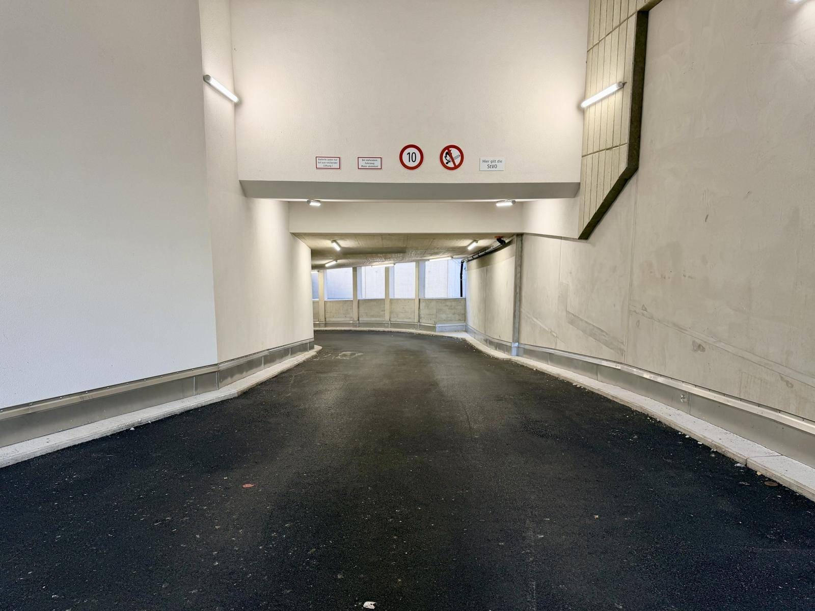 Helle Zufahrtsrampe zu einer modernen Tiefgarage mit Betonwänden und Beleuchtung.