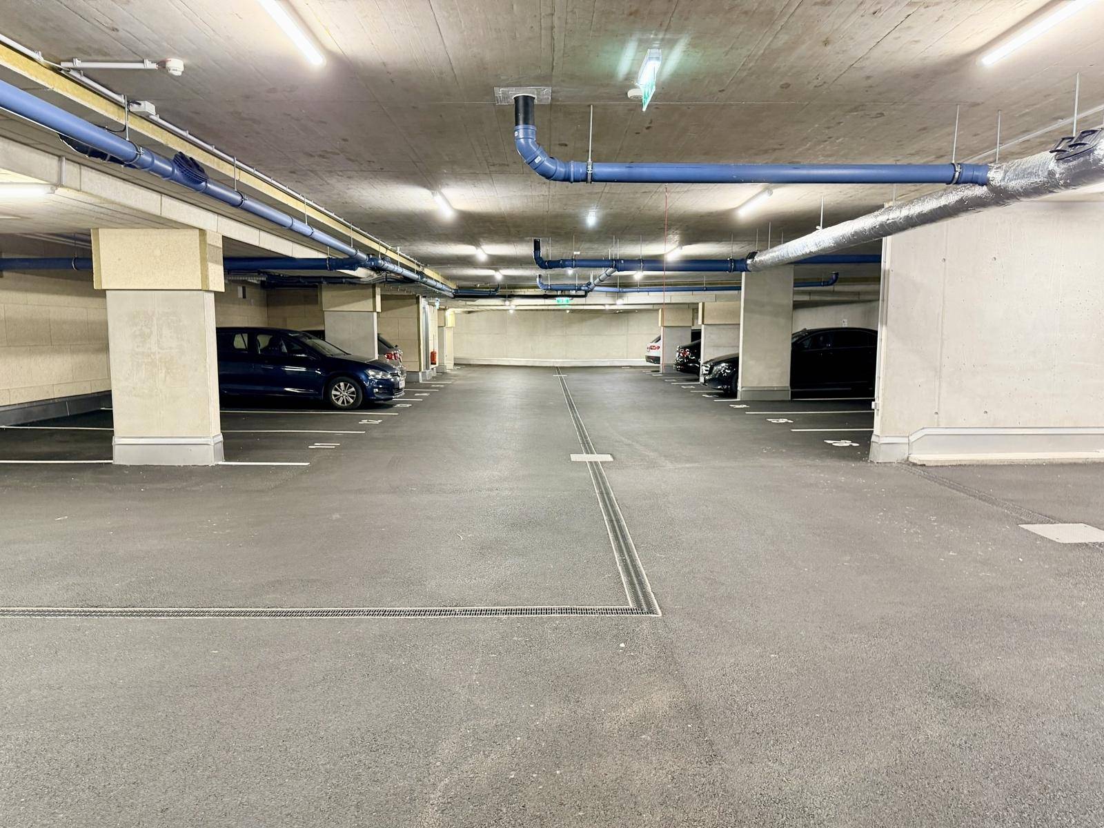 Geräumige Tiefgarage mit mehreren Stellplätzen, Betonpfeilern und sichtbaren Installationen an der Decke.