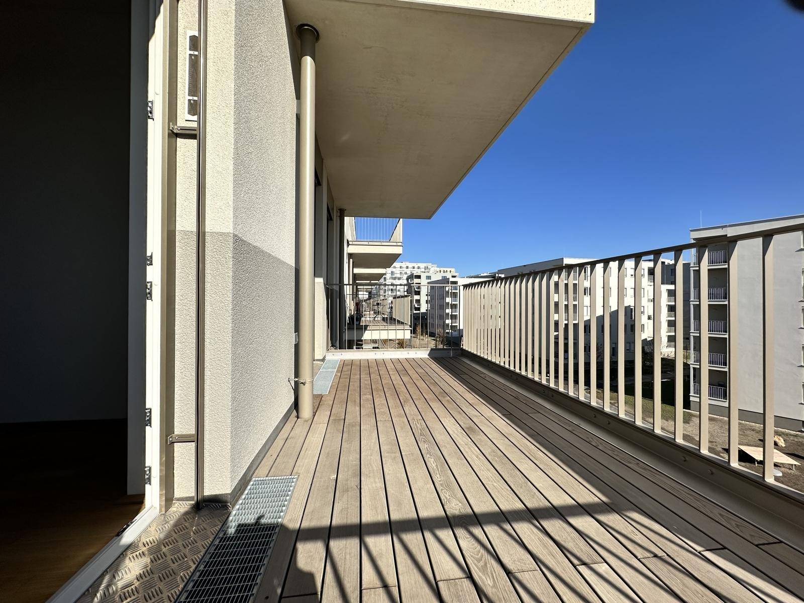 Langer Balkon mit Holzboden und Blick auf die moderne Wohnanlage, sonnig und offen.