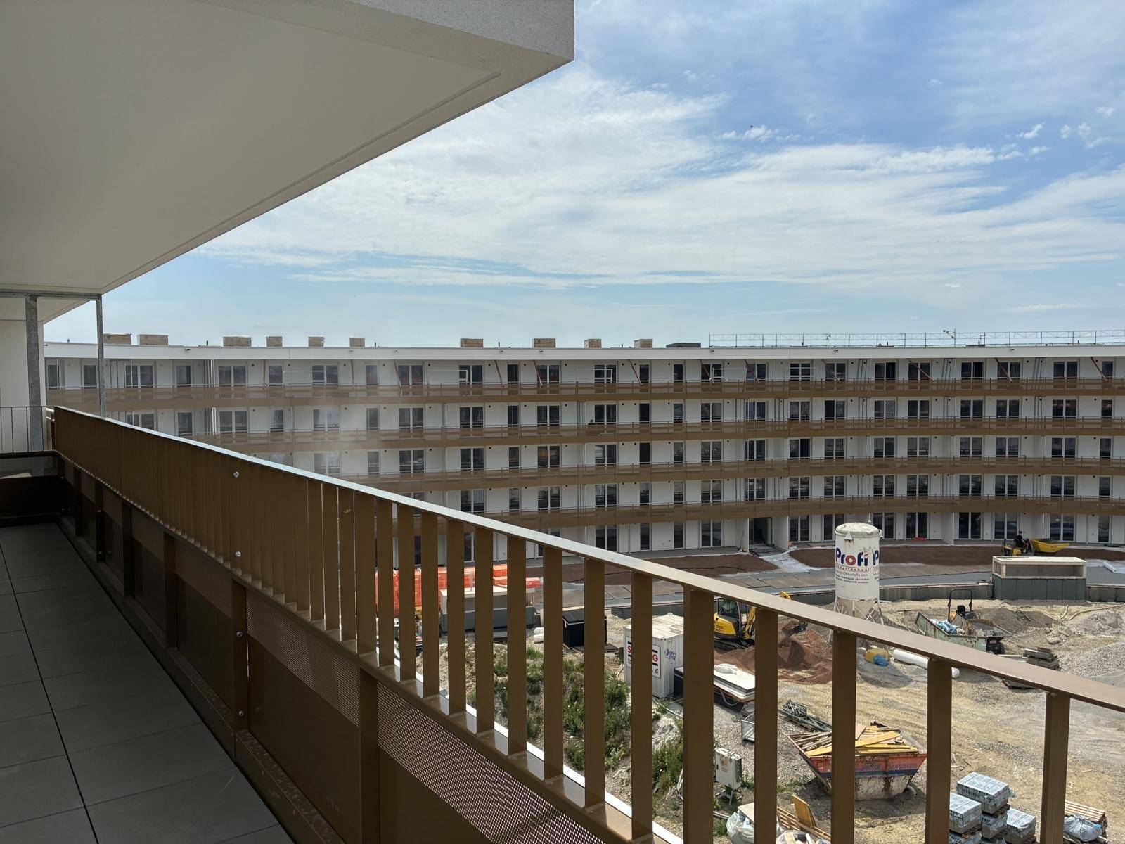 Balkon mit Fliesenboden und Blick auf den im Bau befindlichen Gebäudekomplex unter blauem Himmel.