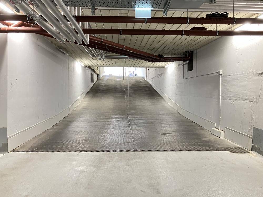 Lange Rampe führt hinab in die Tiefgarage mit sichtbaren Versorgungsleitungen an der Decke.