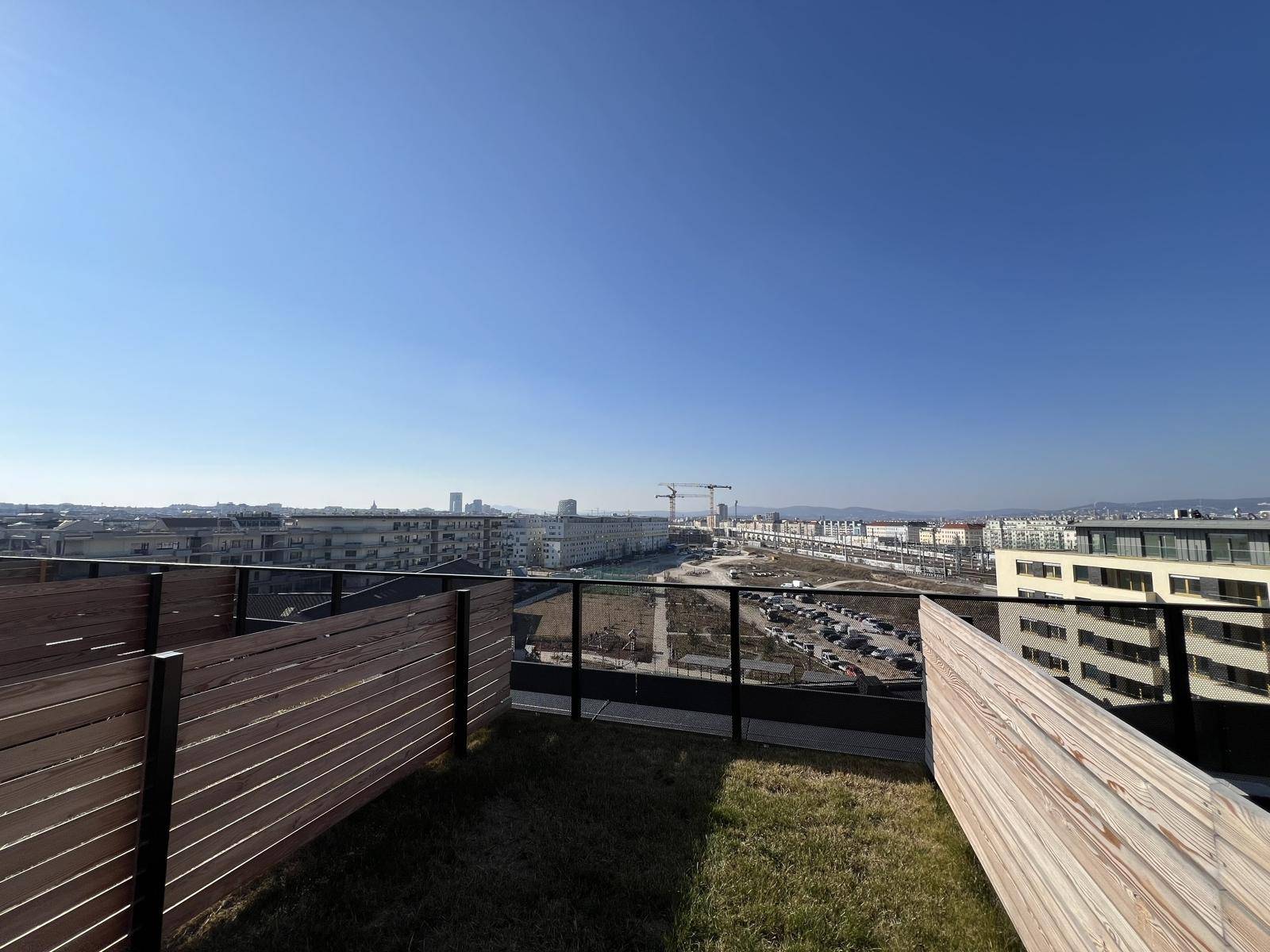 Weitläufiger Stadtblick von einer Dachterrasse mit Grasfläche und niedrigen Holzzäunen im Vordergrund.