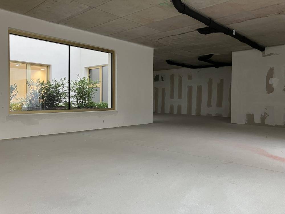 Großer, leerer Raum im Rohbau mit Blick auf einen begrünten Innenhof durch ein breites Fenster.