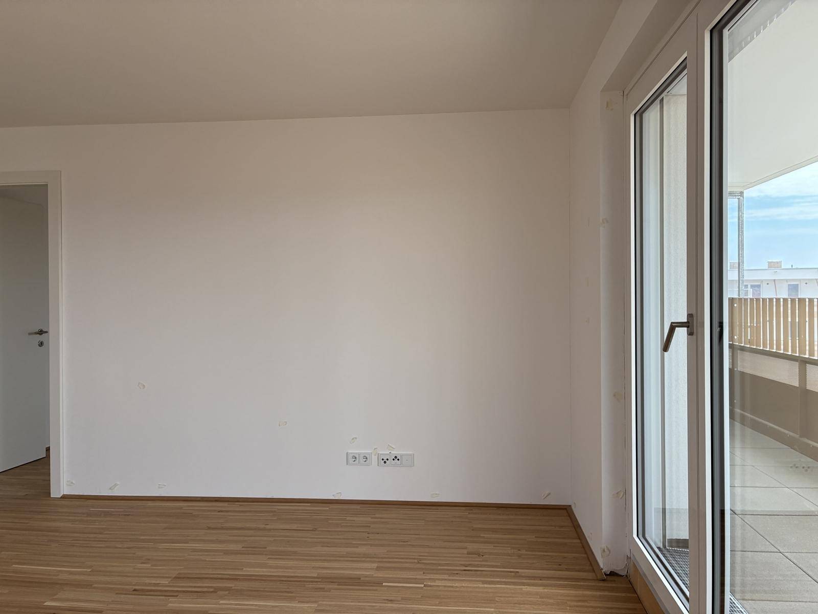 Heller Wohnraum mit Holzboden und großer Fenstertür, die auf einen Balkon führt.