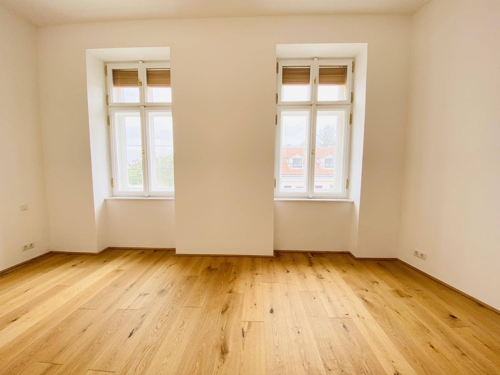 Großes Zimmer mit zwei Fenstern, Holzboden und viel Tageslicht.