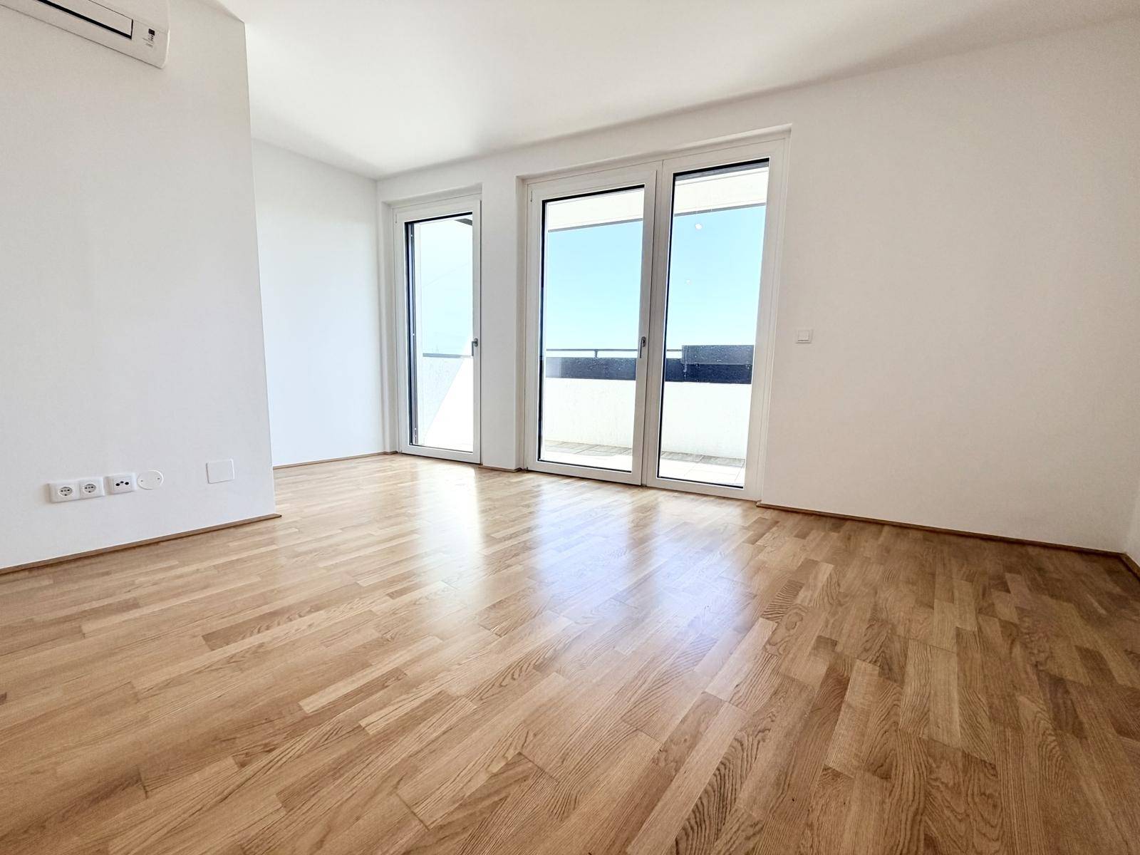 Lichtdurchfluteter Wohnbereich mit Holzboden und großen Fenstern zum Balkon.
