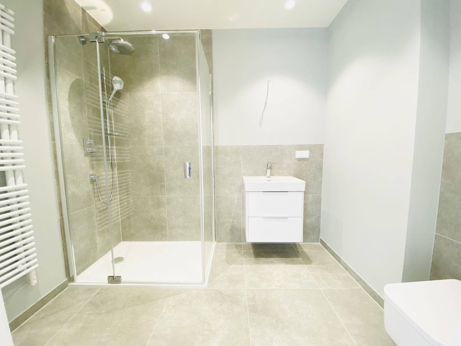 Modernes Badezimmer mit grauen Fliesen, Glasdusche, Regenbrause und beheiztem Handtuchhalter.