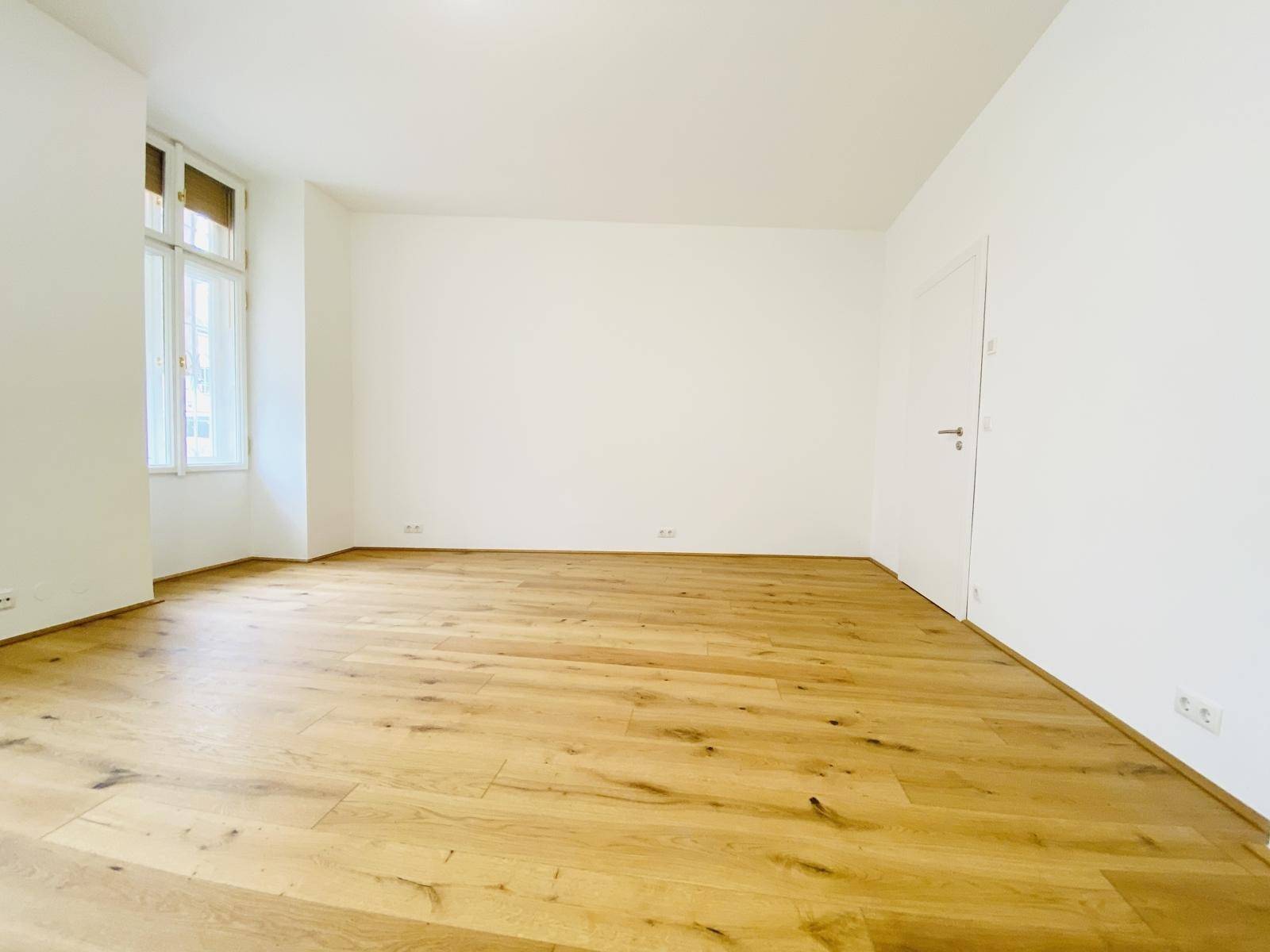 Geräumiges, leeres Zimmer mit hellem Holzboden und großen Fenstern.