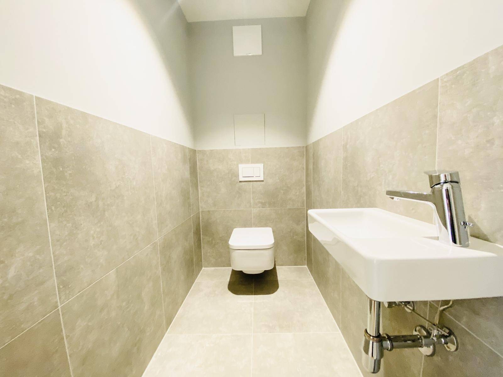 Modernes Gäste-WC mit Wand-WC, kleinem Waschbecken und hellgrauen Fliesen.