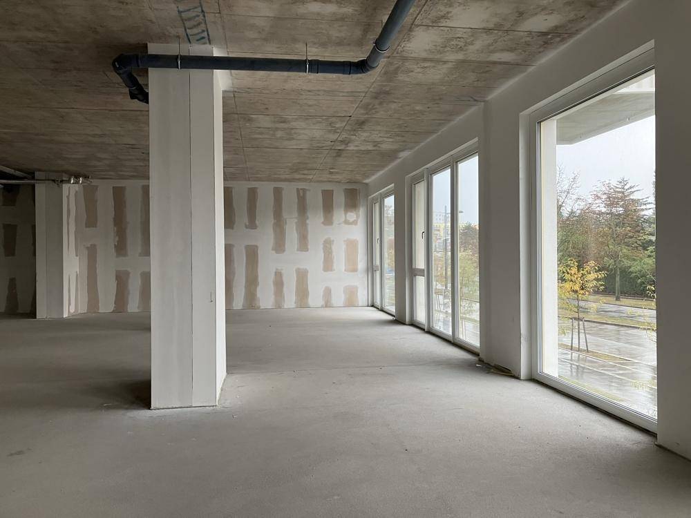 Heller Innenraum im Rohbau mit großen Fensterfronten und sichtbaren Installationen an der Betondecke.
