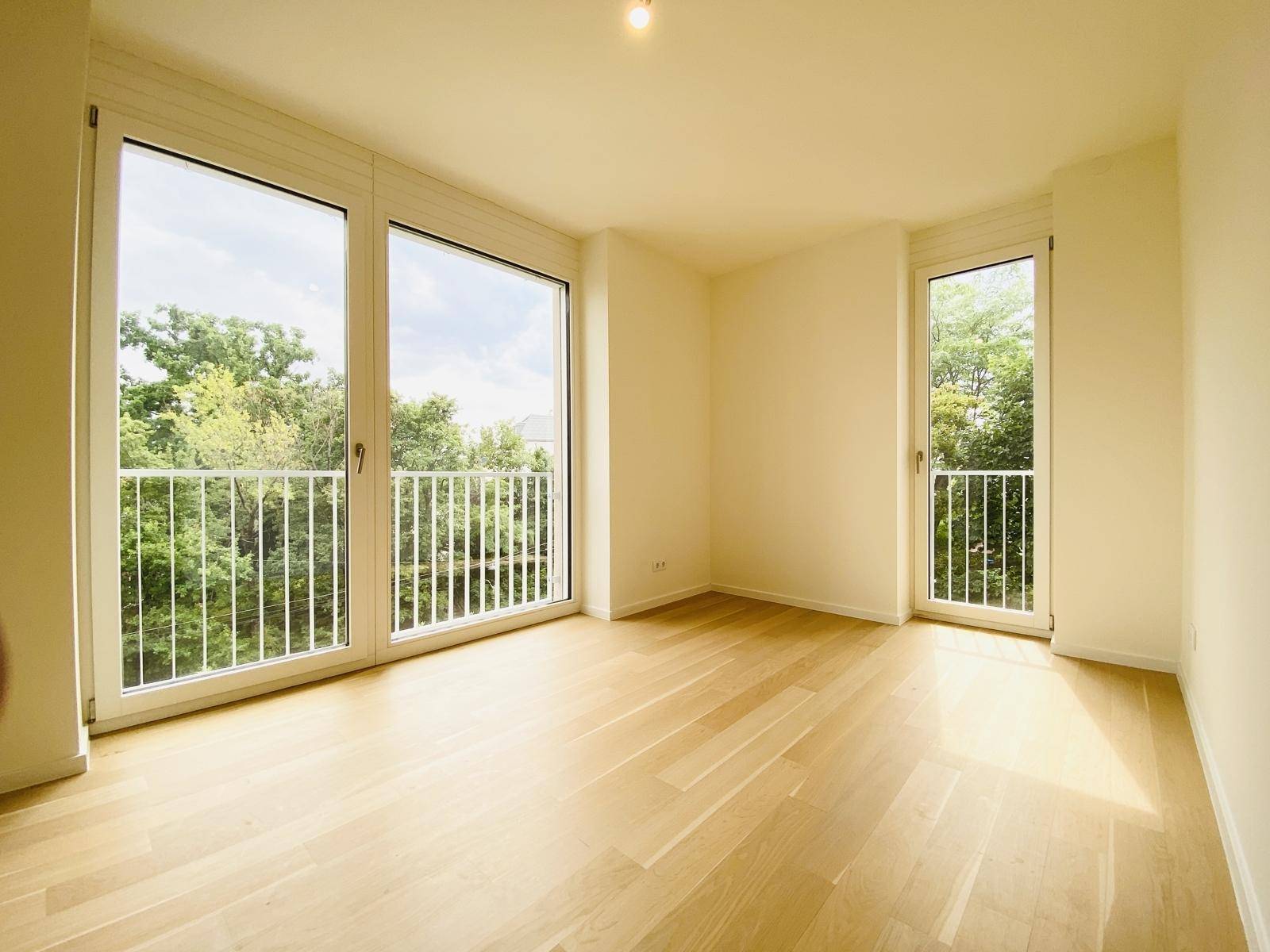 Helles, leeres Zimmer mit großen Fenstern, Parkettboden und Blick auf die grüne Umgebung.