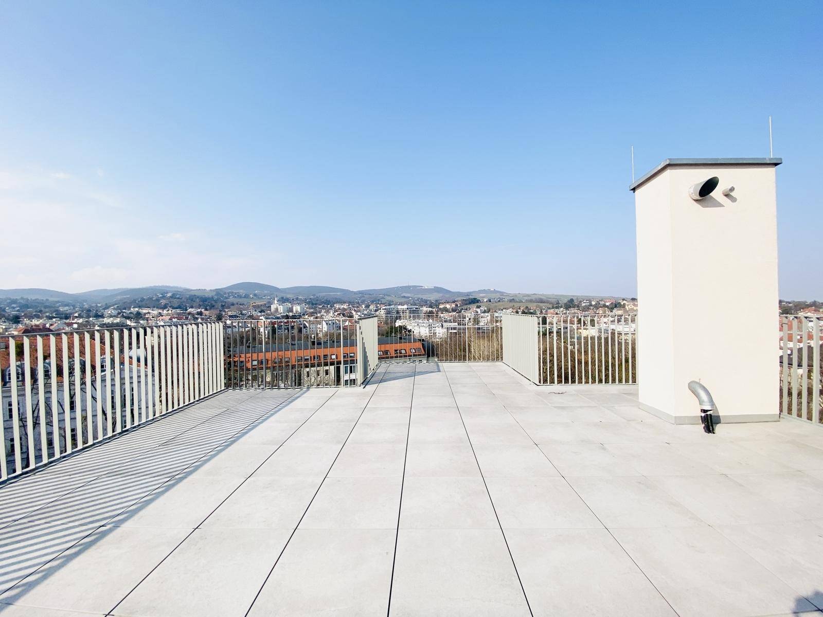 Geräumige Dachterrasse mit Fliesenboden und weitem Blick über die Stadt unter blauem Himmel.
