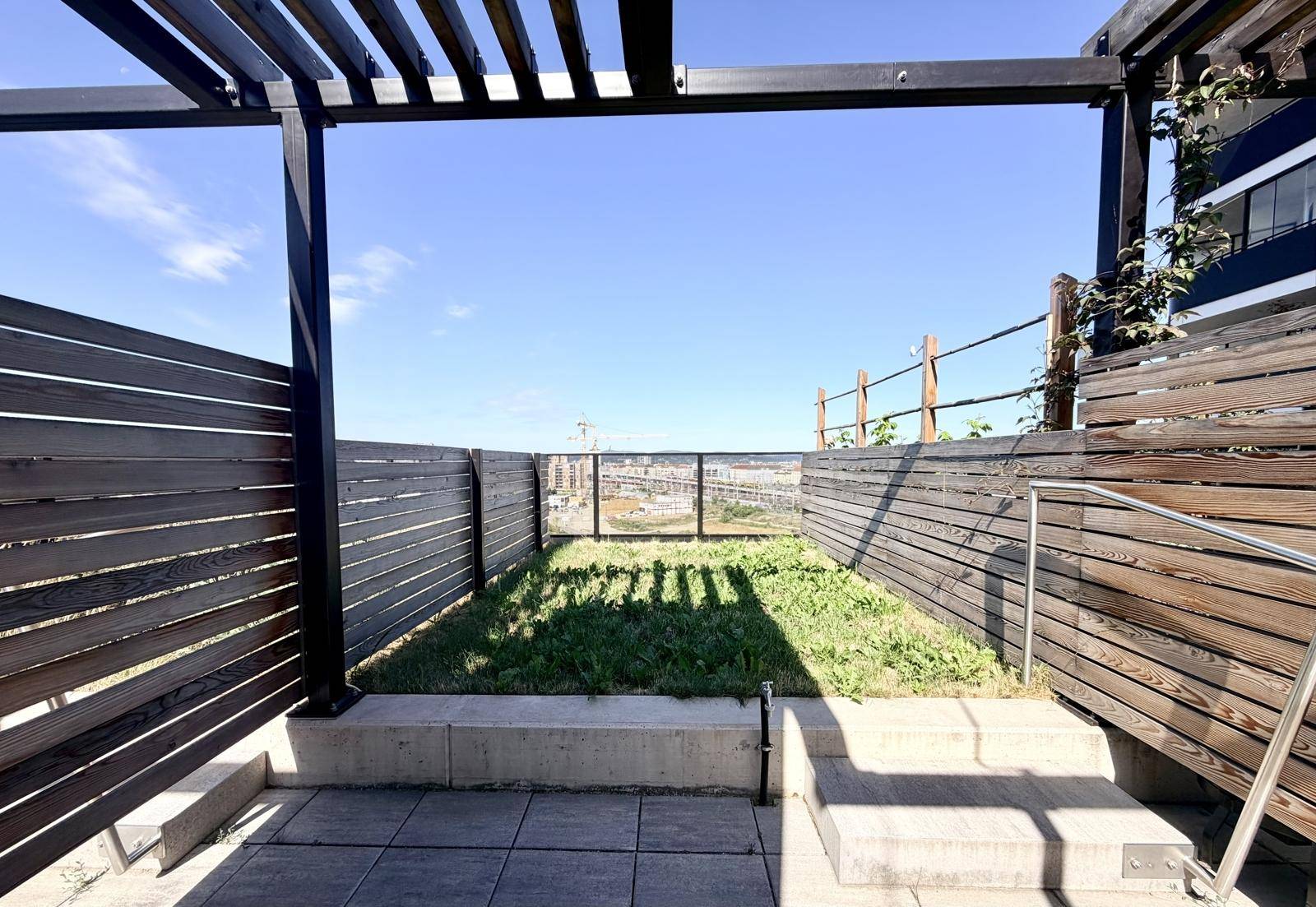 Kleiner Dachgarten mit Rasenfläche, Holzzäunen und Pergola, bietet einen Blick auf die Stadt.