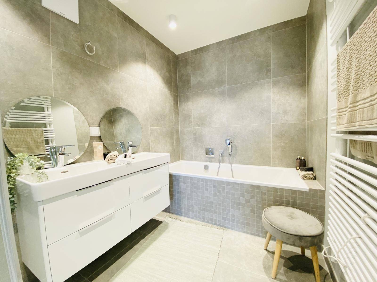 Modernes Badezimmer mit Doppelwaschbecken, Badewanne und beheiztem Handtuchhalter auf grauen Fliesen.