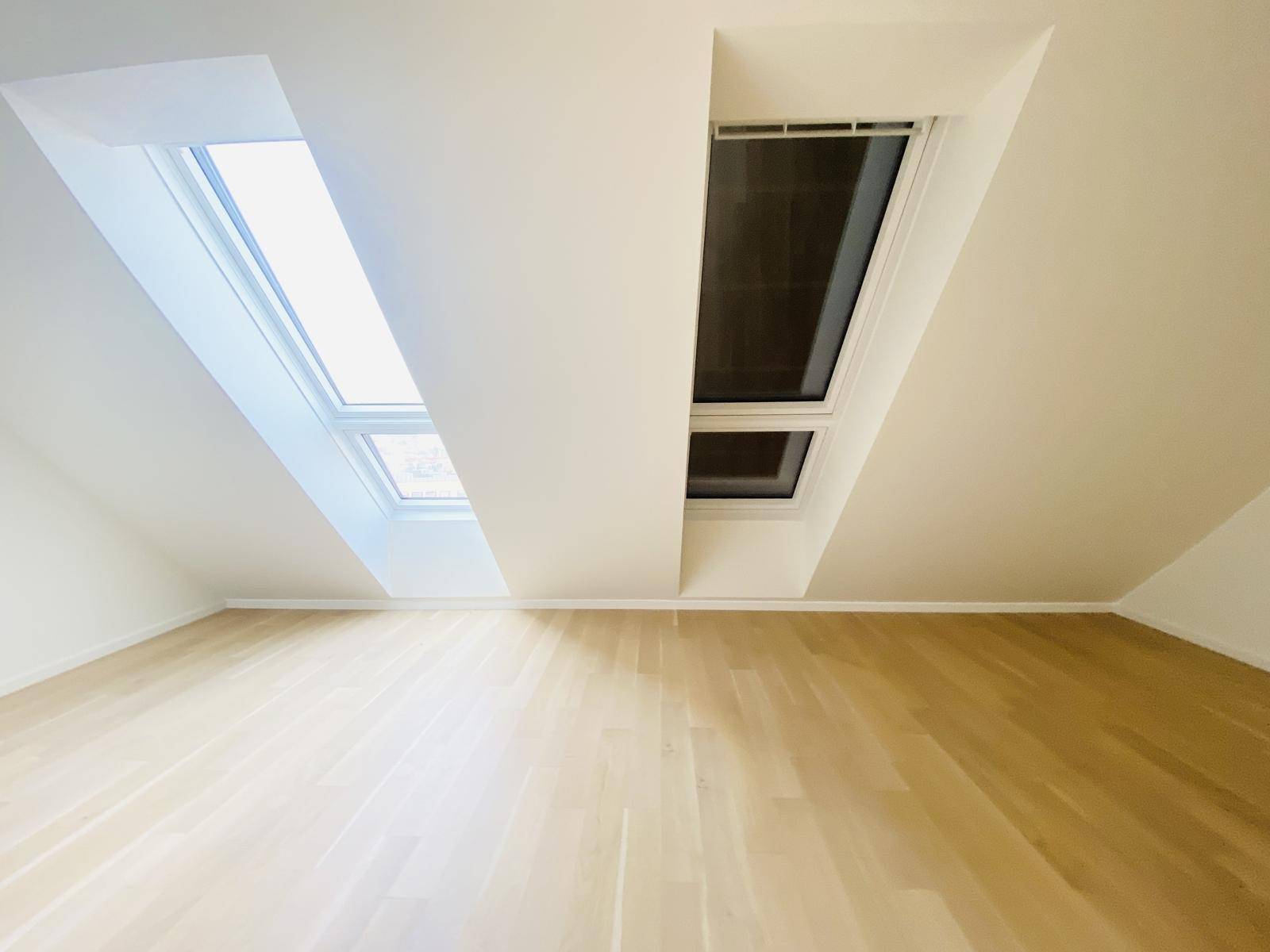 Dachgeschosszimmer mit schrägen Wänden, Holzparkettboden und zwei großen Dachfenstern.