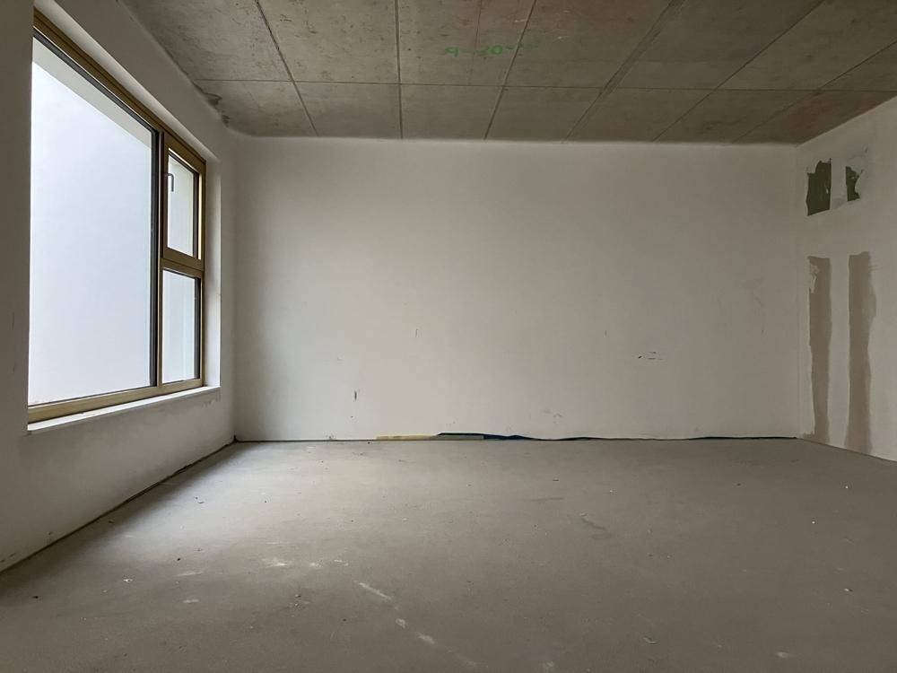 Leerer Raum im Rohbau mit einem großen Fenster, das viel Tageslicht hereinlässt und eine helle Atmosphäre schafft.