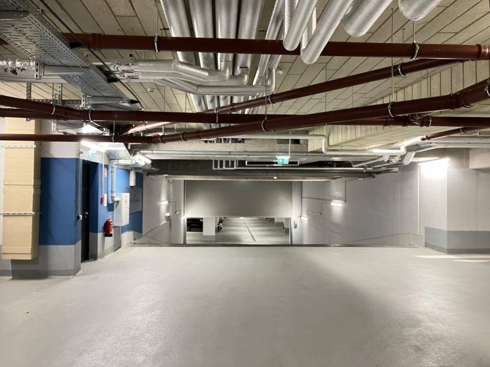 Heller Bereich in der Tiefgarage mit sichtbaren Leitungen und Zugang zu weiteren Parkebenen.