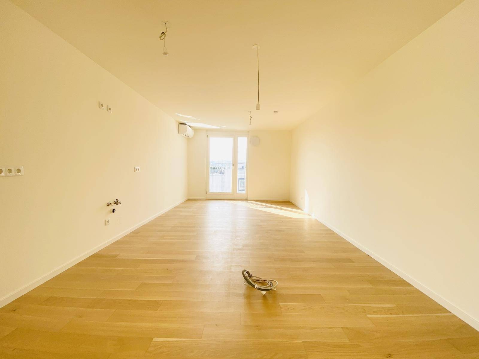 Geräumiges, leeres Wohnzimmer mit hellem Holzparkettboden und Zugang zu einem Balkon.