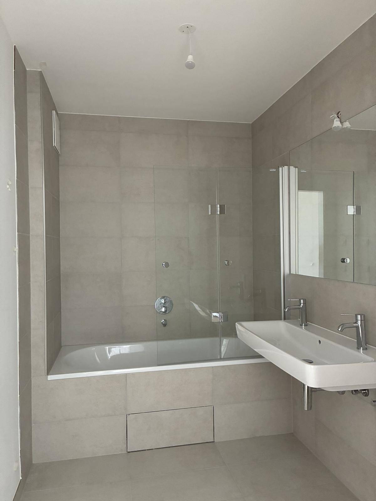 Modernes Badezimmer mit hellgrauen Fliesen, Badewanne mit Duschwand und Einzelwaschbecken.