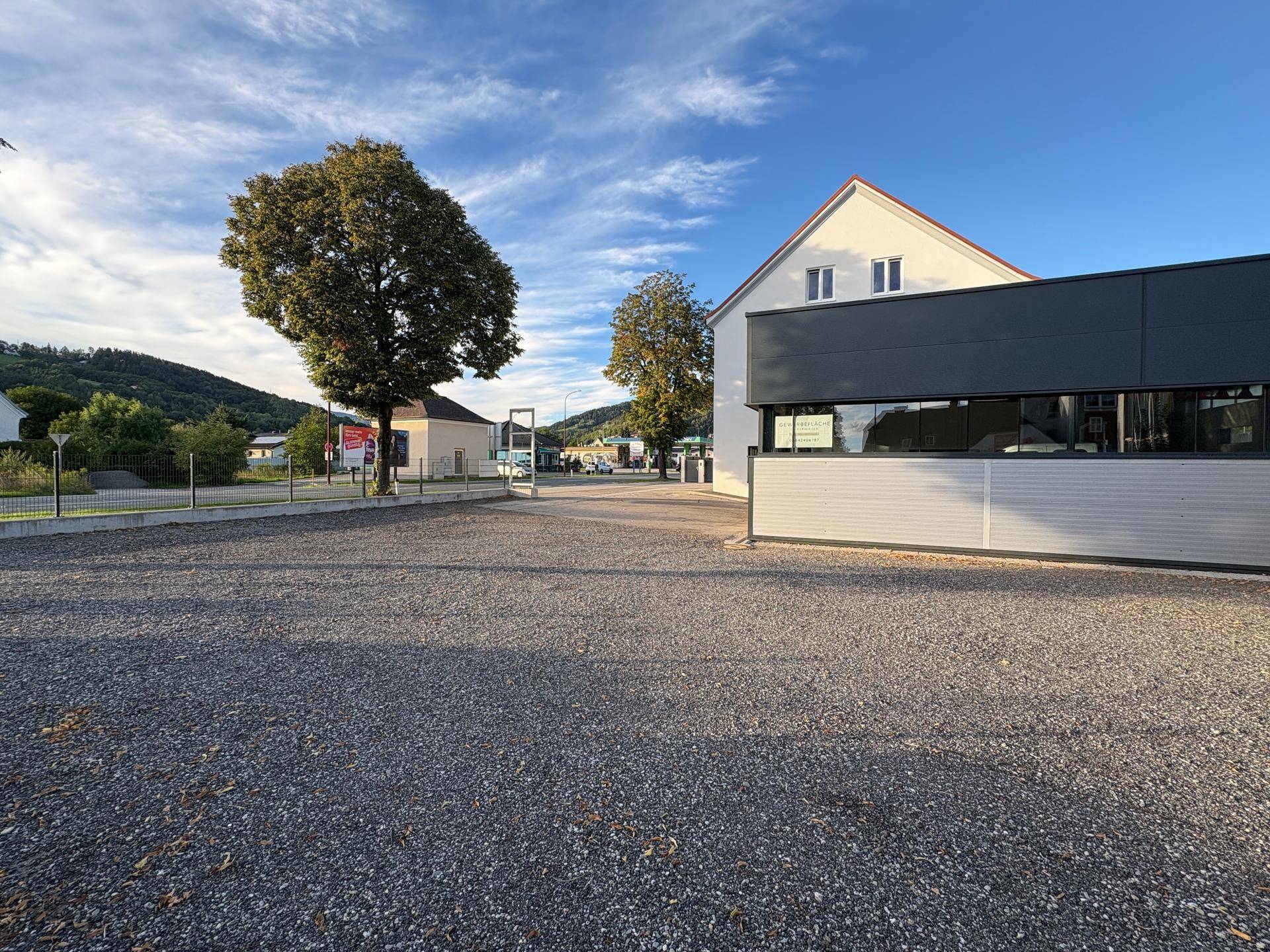 Weitläufiger Schotterparkplatz vor dem Gewerbegebäude mit Bäumen und Hügeln im Hintergrund.