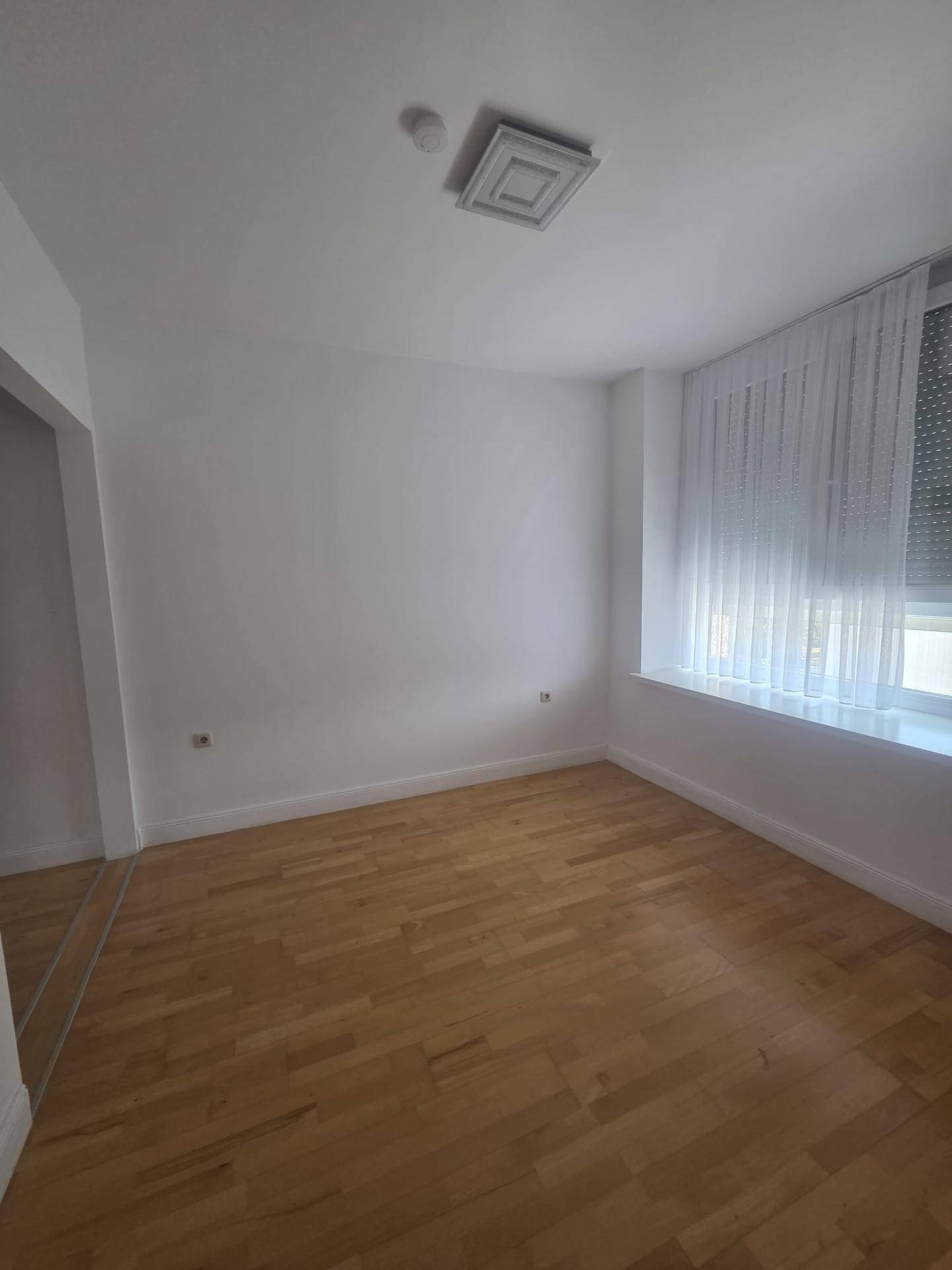 Helles Zimmer mit Holzboden, weißen Wänden und einem Fenster mit Jalousien.